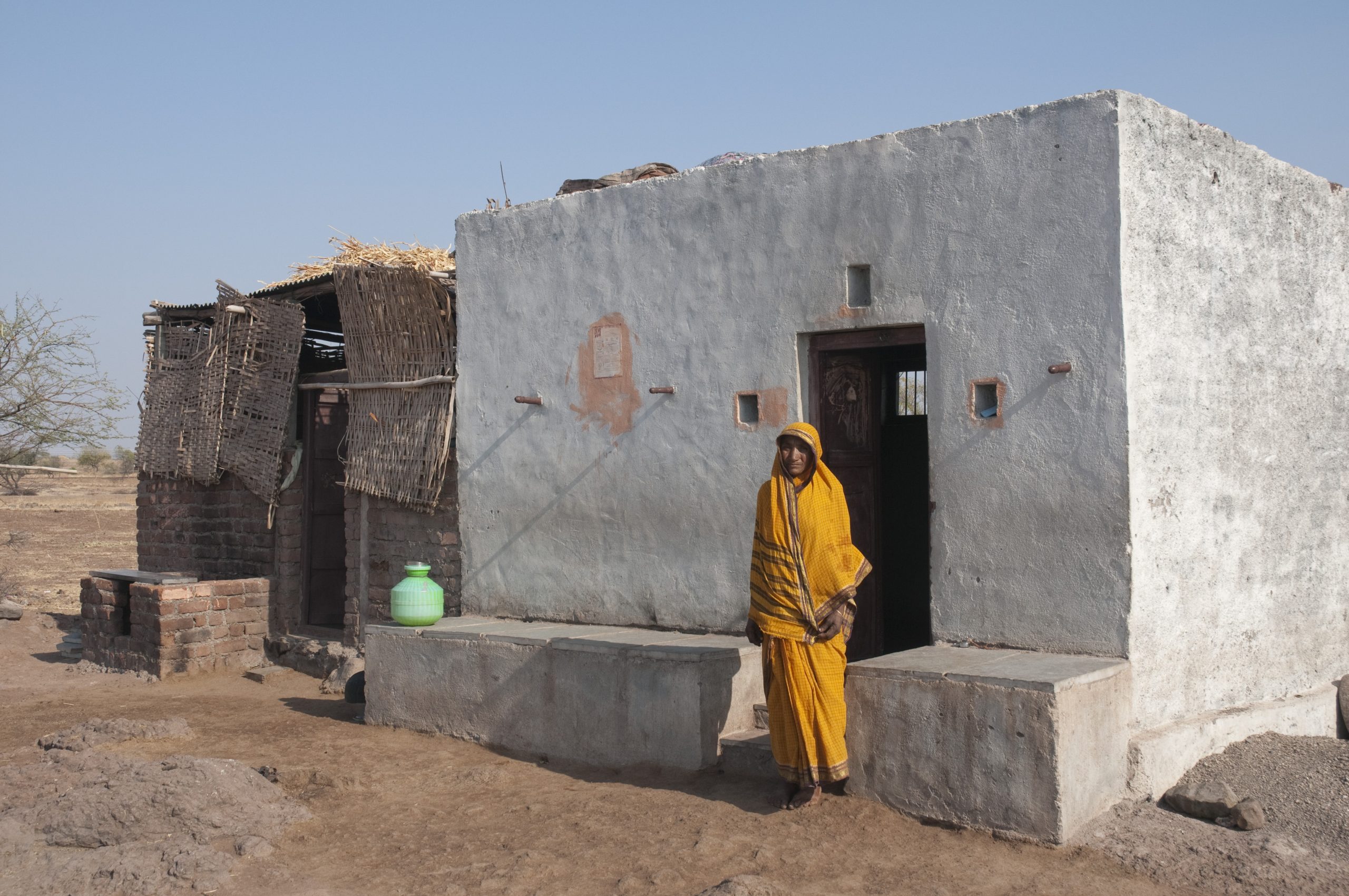 Een vrouw in een geel gewaad staat voor een betonnen huis in India.