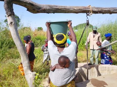 Een vrouw met een teil staat bij een waterput.